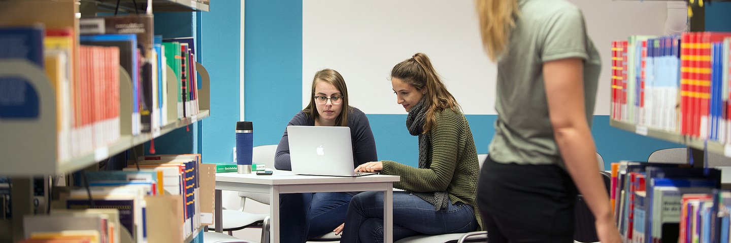 Zwei Personen am Laptop am Tisch, eine Person sucht ein Buch aus einem Regal raus