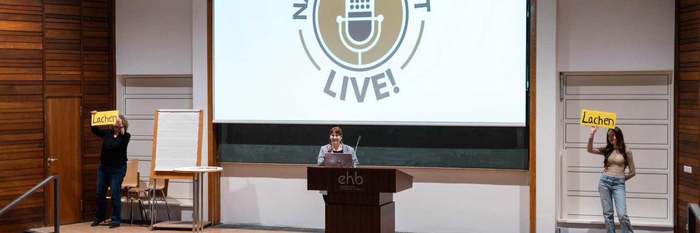 drei Studierende stehen auf einer bühne vor einem projektor, auf dem „nachsbarschaft live“ steht. einer von ihnen präsentiert hinter einem podium, während die beiden anderen schilder mit der Aufschrift „lachen“ hochhalten